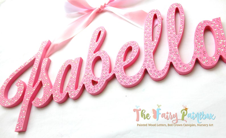 May include: Pink wooden letters spelling the name "Isabella" with small white rhinestones embedded in the paint. The letters are in a cursive font and are hanging from a pink ribbon.