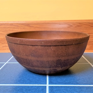 May include: A large, round, brown mixing bowl. The bowl is made of a dark brown material and has a wide rim. It sits on a blue tiled surface with white grout. The background features a wooden ledge and a yellow wall.