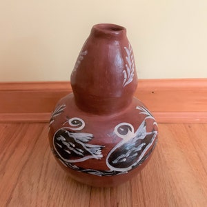 May include: A handcrafted, brown gourd-shaped vase with a narrow neck. The vase features white painted designs of birds and foliage. The gourd is sitting on a wooden surface.