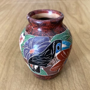 May include: A small, round ceramic vase with a textured, reddish-brown exterior. The vase features a painted design of a toucan bird and colorful flowers in shades of purple, green, and red. The rim of the vase is a darker shade of brown.
