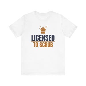 以下が含まれることがあります： 白い半袖Tシャツで、上に泡がある茶色のバケツのグラフィックが描かれています。グラフィックの下には、濃い青色の「LICENSED」と茶色の「TO SCRUB」という文字があり、2つのフレーズの間に水平線が引かれています。
