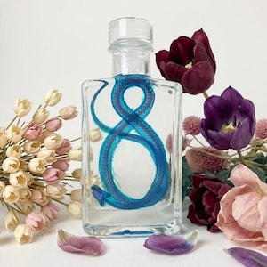 May include: A clear glass rectangular bottle containing a vibrant blue snake skeleton suspended in liquid. The bottle is surrounded by a cluster of dried cream-colored flowers on the left and deep purple tulips on the right.