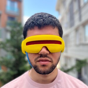 May include: A bright yellow visor with a horizontal red mesh lens covering the eyes. The eyewear features a smooth, curved frame design that extends across the forehead and temples, secured by a blue strap visible on the side of the head.