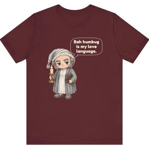 Puede incluir: Una camiseta de manga corta color granate con una ilustraci&oacute;n de dibujos animados de un personaje con gorro de dormir y bata que sostiene una vela y un libro. Un bocadillo contiene el texto: Bah humbug is my love language.