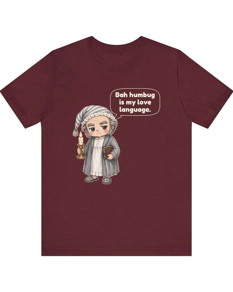 Puede incluir: Una camiseta de manga corta color granate con una ilustraci&oacute;n de dibujos animados de un personaje con gorro de dormir y bata que sostiene una vela y un libro. Un bocadillo contiene el texto: Bah humbug is my love language.