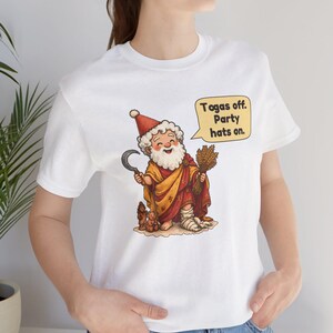 May include: A white t-shirt featuring a cartoon illustration of a bearded figure wearing a red hat and yellow robe, holding a sickle and wheat. A speech bubble contains the text: Togas off. Party hats on. The figure has a bandaged foot.