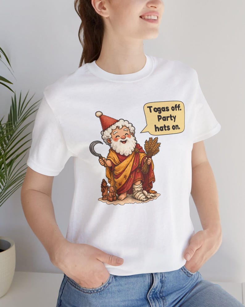 May include: A white t-shirt featuring a cartoon illustration of a bearded figure wearing a red hat and yellow robe, holding a sickle and wheat. A speech bubble contains the text: Togas off. Party hats on. The figure has a bandaged foot.