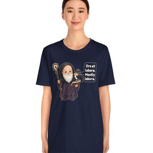 May include: A navy blue t-shirt featuring a cartoon illustration of a bearded monk in a dark robe holding a wooden staff and an open book. A black raven sits on the monk's shoulder. The text reads: Ora et labora. Mostly labora.