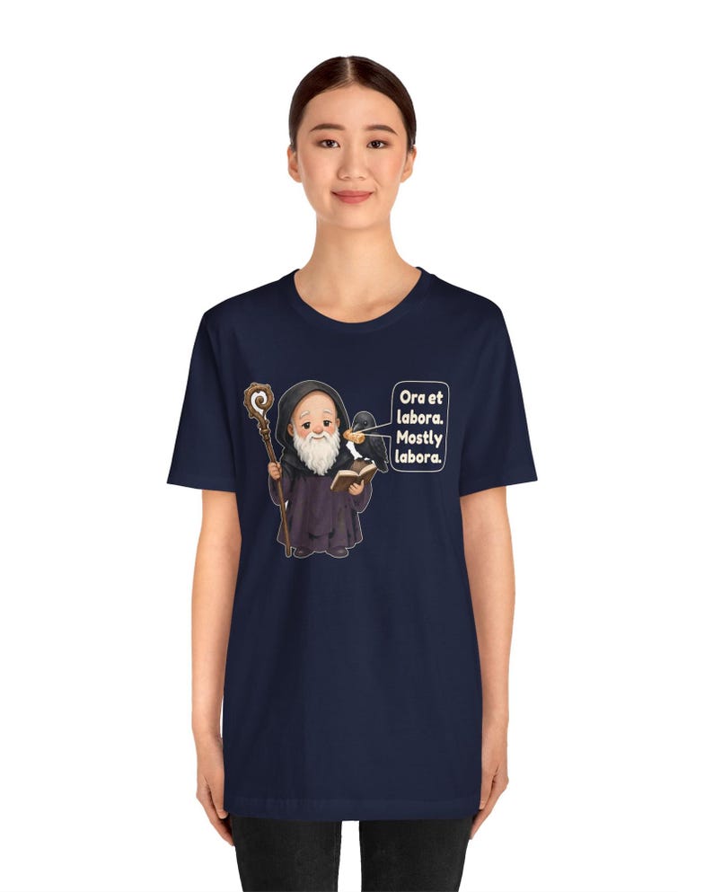 May include: A navy blue t-shirt featuring a cartoon illustration of a bearded monk in a dark robe holding a wooden staff and an open book. A black raven sits on the monk's shoulder. The text reads: Ora et labora. Mostly labora.