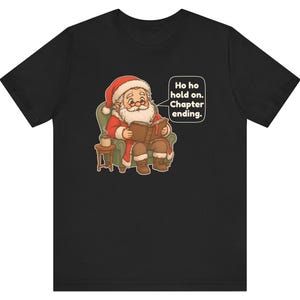 Op de afbeelding: Een zwart t-shirt met een afbeelding van Santa Claus die in een groene fauteuil zit en een boek leest. Naast hem staat een klein tafeltje met een mok. Een tekstballon bevat de tekst: Ho ho hold on. Chapter ending. Het ontwerp is gecentreerd.