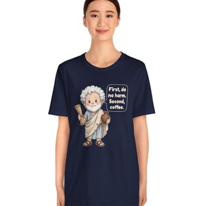 May include: A navy blue t-shirt featuring a cartoon illustration of a white-haired man in a light blue toga holding a scroll and a brown mug. A speech bubble next to him contains the white text: First, do no harm. Second, coffee.