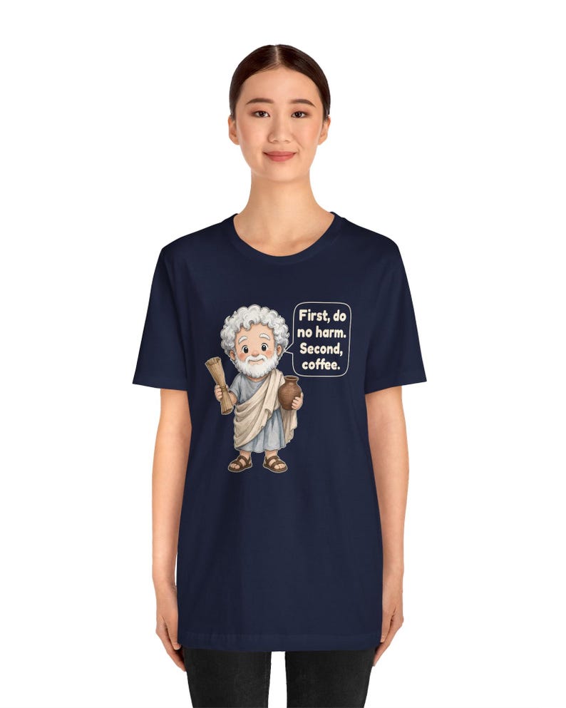 May include: A navy blue t-shirt featuring a cartoon illustration of a white-haired man in a light blue toga holding a scroll and a brown mug. A speech bubble next to him contains the white text: First, do no harm. Second, coffee.