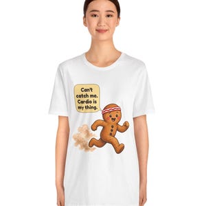 May include: A white short-sleeve t-shirt featuring a graphic of a brown gingerbread person wearing a red and white headband, running and kicking up dust. A yellow speech bubble contains the text: Can't catch me. Cardio is my thing.