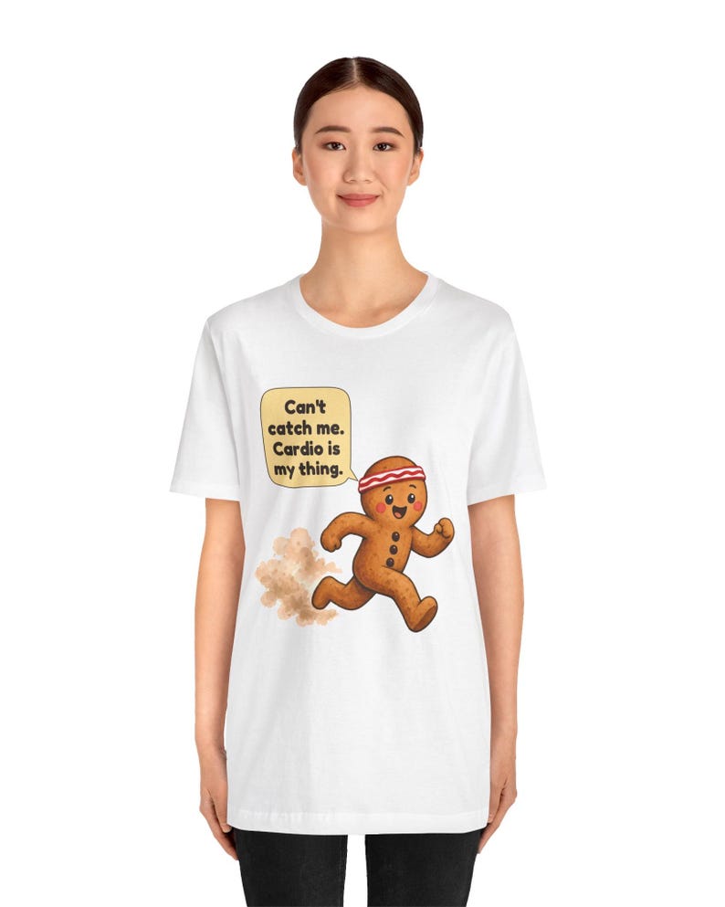 May include: A white short-sleeve t-shirt featuring a graphic of a brown gingerbread person wearing a red and white headband, running and kicking up dust. A yellow speech bubble contains the text: Can't catch me. Cardio is my thing.