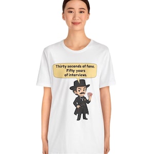 Pu&ograve; includere: Una t-shirt bianca a maniche corte con un'illustrazione a cartoni animati di un uomo in abito nero e cilindro che tiene in mano delle carte da gioco. Sopra la figura c'&egrave; un fumetto giallo con il testo: Thirty seconds of fame. Fifty years of interviews.
