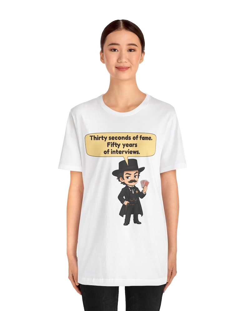 Pu&ograve; includere: Una t-shirt bianca a maniche corte con un'illustrazione a cartoni animati di un uomo in abito nero e cilindro che tiene in mano delle carte da gioco. Sopra la figura c'&egrave; un fumetto giallo con il testo: Thirty seconds of fame. Fifty years of interviews.