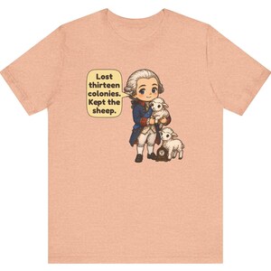 Peut inclure: Un t-shirt couleur p&ecirc;che avec une illustration de style dessin anim&eacute; repr&eacute;sentant un personnage historique en manteau bleu tenant un mouton blanc, accompagn&eacute; d'un autre mouton et d'une petite horloge. Une bulle contient le texte : Lost thirteen colonies. Kept the sheep.