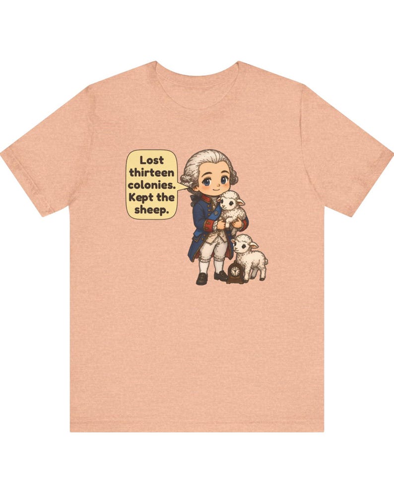 Peut inclure: Un t-shirt couleur p&ecirc;che avec une illustration de style dessin anim&eacute; repr&eacute;sentant un personnage historique en manteau bleu tenant un mouton blanc, accompagn&eacute; d'un autre mouton et d'une petite horloge. Une bulle contient le texte : Lost thirteen colonies. Kept the sheep.