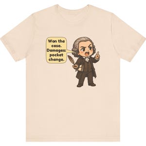 May include: A light beige t-shirt featuring a cartoon illustration of a historical figure in a brown suit holding a quill. A speech bubble next to the character contains the text: Won the case. Damages: pocket change.