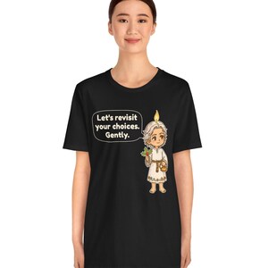 May include: A black t-shirt featuring a graphic of a small, light-haired figure with a candle flame on their head, holding a sprig of holly and a small basket. A speech bubble next to the figure contains the text, Let's revisit your choices. Gently.