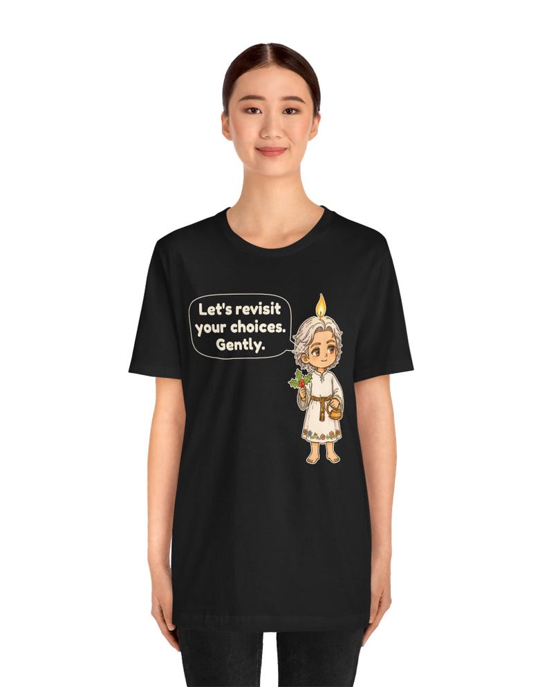 May include: A black t-shirt featuring a graphic of a small, light-haired figure with a candle flame on their head, holding a sprig of holly and a small basket. A speech bubble next to the figure contains the text, Let's revisit your choices. Gently.