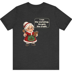 K&ouml;nnte beinhalten: Ein dunkelgraues T-Shirt mit kurzen &Auml;rmeln und einer Grafik einer Frau in einem rot-wei&szlig;en Weihnachtskost&uuml;m mit gr&uuml;ner Sch&uuml;rze, die einen Holzl&ouml;ffel und ein Klemmbrett h&auml;lt, mit dem Text: I run the workshop. He gets the credit.