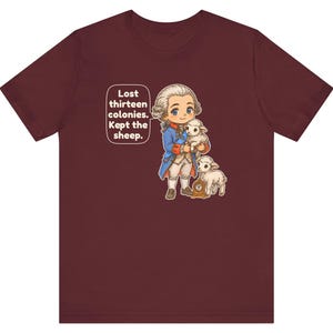 Peut inclure: Un t-shirt &agrave; manches courtes bordeaux avec une illustration de style dessin anim&eacute; repr&eacute;sentant un personnage historique en manteau bleu tenant un agneau blanc, avec un autre agneau et une petite horloge. Texte : Lost thirteen colonies. Kept the sheep.