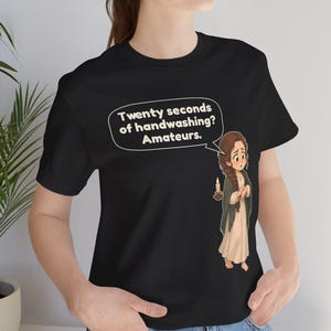 Op de afbeelding: Een zwart t-shirt met korte mouwen en een cartoonillustratie van een persoon met een lange vlecht die een kaars vasthoudt. Een tekstballon boven het personage bevat de tekst: Twenty seconds of handwashing? Amateurs.
