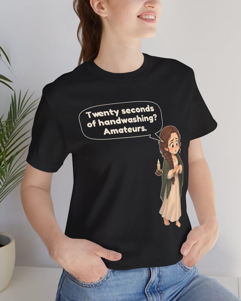 Op de afbeelding: Een zwart t-shirt met korte mouwen en een cartoonillustratie van een persoon met een lange vlecht die een kaars vasthoudt. Een tekstballon boven het personage bevat de tekst: Twenty seconds of handwashing? Amateurs.