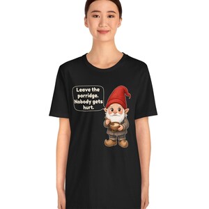 May include: A black short-sleeved t-shirt featuring a graphic of a gnome wearing a red hat and holding a bowl of porridge. A speech bubble next to the gnome contains the text: Leave the porridge. Nobody gets hurt.