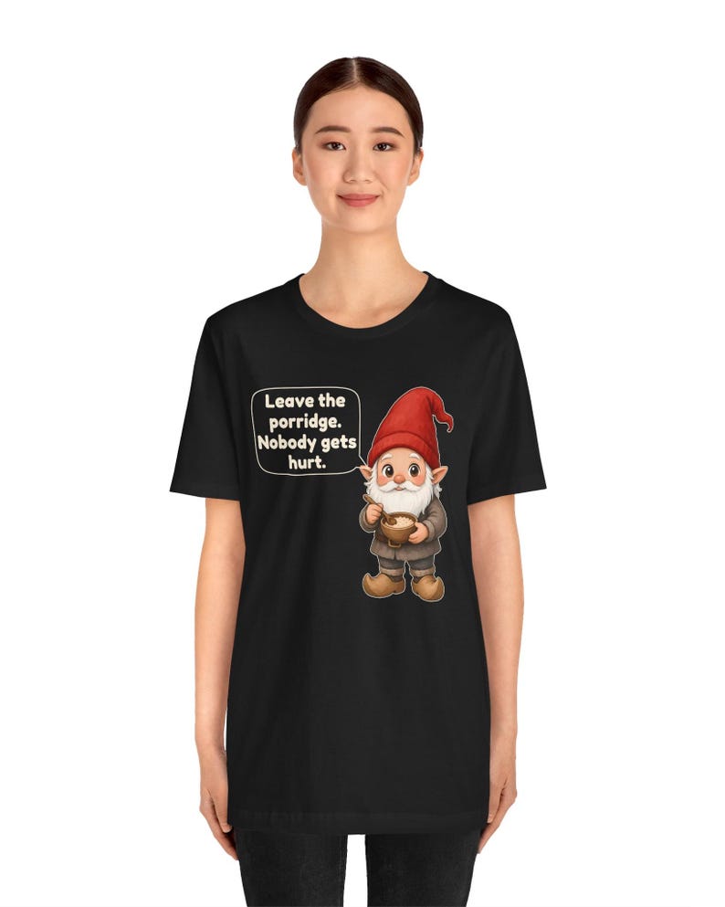 May include: A black short-sleeved t-shirt featuring a graphic of a gnome wearing a red hat and holding a bowl of porridge. A speech bubble next to the gnome contains the text: Leave the porridge. Nobody gets hurt.