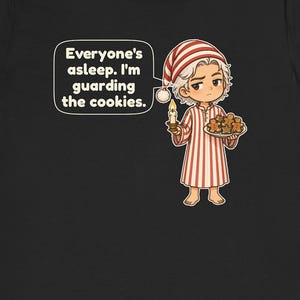 Op de afbeelding: Een zwart t-shirt met een stripfiguur in een rood-wit gestreepte pyjama en een bijpassende slaapmuts. Het figuur houdt een brandende kaars en een bord met peperkoekkoekjes vast. Een tekstballon bevat de tekst: Everyone's asleep. I'm guarding the cookies.