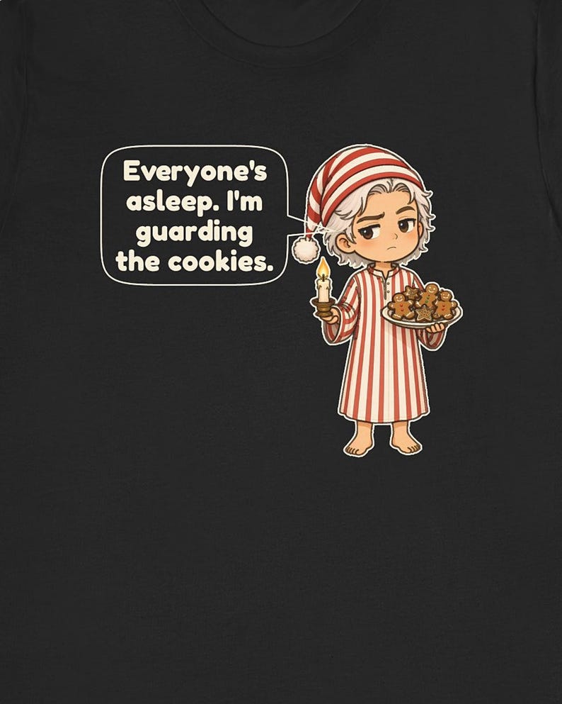 Op de afbeelding: Een zwart t-shirt met een stripfiguur in een rood-wit gestreepte pyjama en een bijpassende slaapmuts. Het figuur houdt een brandende kaars en een bord met peperkoekkoekjes vast. Een tekstballon bevat de tekst: Everyone's asleep. I'm guarding the cookies.