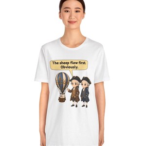 May include: A white t-shirt featuring a graphic of two historical figures in 18th-century attire standing next to a hot air balloon with a sheep inside. A speech bubble above them contains the text: The sheep flew first. Obviously.