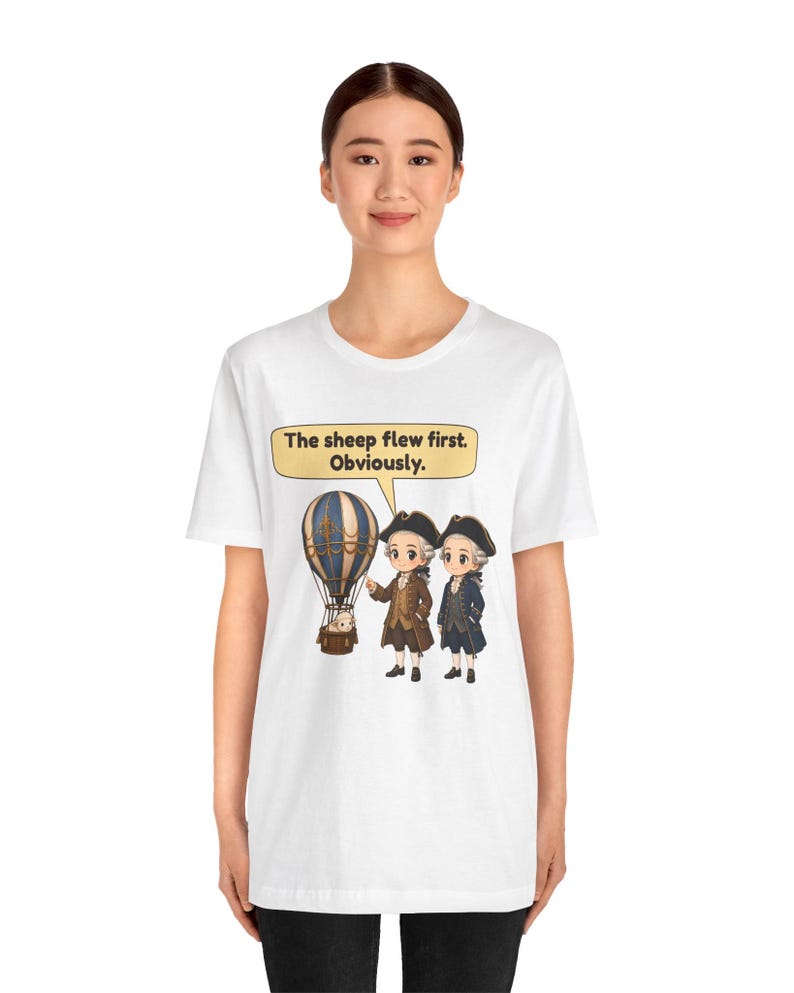 May include: A white t-shirt featuring a graphic of two historical figures in 18th-century attire standing next to a hot air balloon with a sheep inside. A speech bubble above them contains the text: The sheep flew first. Obviously.