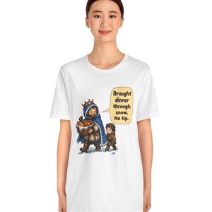 May include: A white t-shirt featuring a graphic of a king in a blue cloak carrying a basket of bread and a small child walking through snow. A speech bubble next to them contains the text: Brought dinner through snow. No tip.