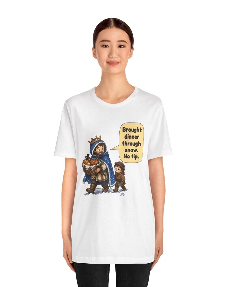 May include: A white t-shirt featuring a graphic of a king in a blue cloak carrying a basket of bread and a small child walking through snow. A speech bubble next to them contains the text: Brought dinner through snow. No tip.