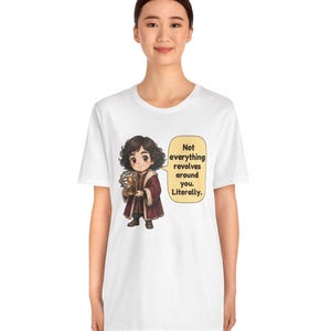 May include: A white short-sleeved t-shirt featuring a cartoon illustration of a person in a dark red coat holding a model of the solar system. A speech bubble next to the character contains the text: Not everything revolves around you. Literally.