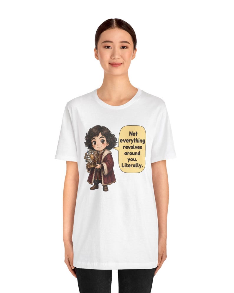 May include: A white short-sleeved t-shirt featuring a cartoon illustration of a person in a dark red coat holding a model of the solar system. A speech bubble next to the character contains the text: Not everything revolves around you. Literally.
