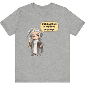 Puede incluir: Una camiseta gris jaspeado con una ilustraci&oacute;n de dibujos animados de un personaje con gorro de dormir y bata que sostiene una vela y un libro. Un globo de texto sobre el personaje contiene: Bah humbug is my love language.