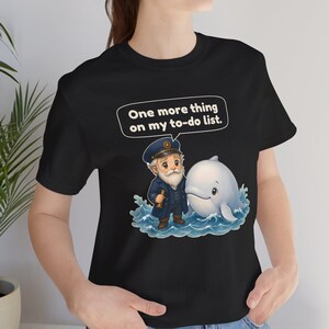 May include: A black t-shirt featuring a graphic of a bearded captain in a navy uniform holding a telescope next to a white whale in blue water. Above the characters, a speech bubble contains the text, One more thing on my to-do list.