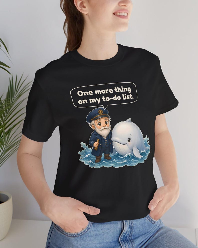 May include: A black t-shirt featuring a graphic of a bearded captain in a navy uniform holding a telescope next to a white whale in blue water. Above the characters, a speech bubble contains the text, One more thing on my to-do list.