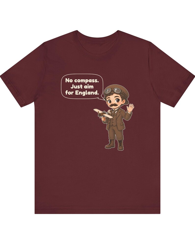 May include: A maroon t-shirt featuring a cartoon illustration of an aviator with a mustache, wearing a brown suit, cap, and goggles. The aviator holds a small airplane and a speech bubble reads: No compass. Just aim for England.