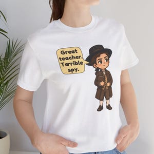 May include: A white t-shirt featuring a cartoon illustration of a person in 18th-century brown attire and a black hat, holding a book. A yellow speech bubble next to the character contains the black text: Great teacher. Terrible spy.