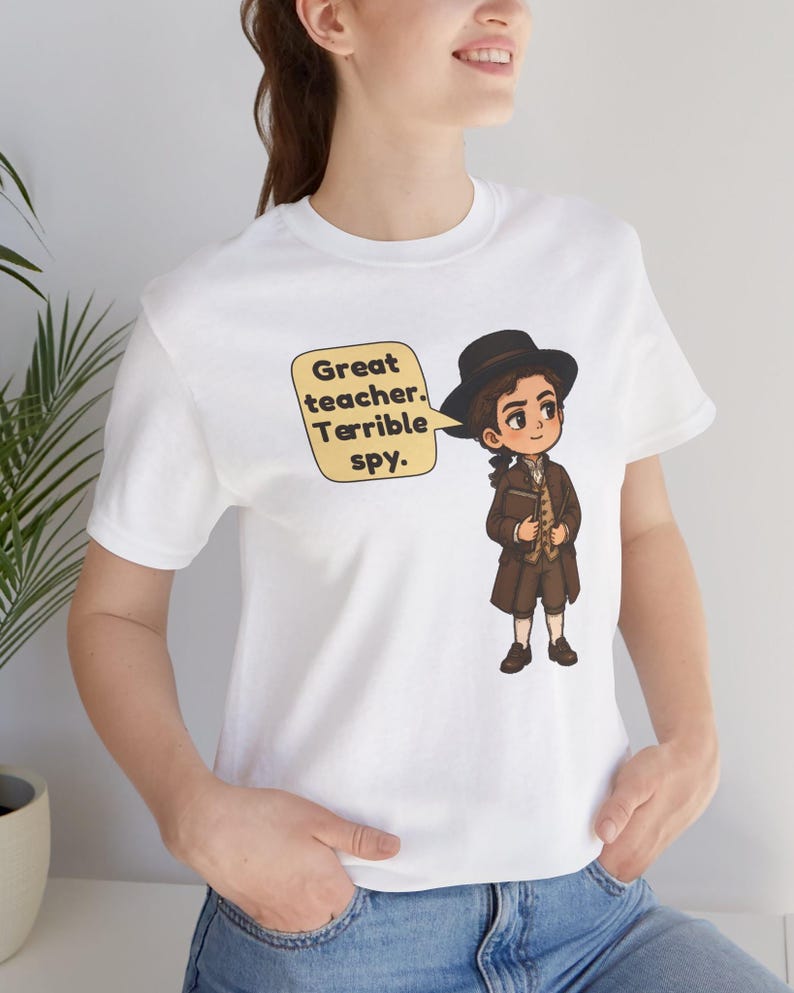 May include: A white t-shirt featuring a cartoon illustration of a person in 18th-century brown attire and a black hat, holding a book. A yellow speech bubble next to the character contains the black text: Great teacher. Terrible spy.