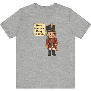 Pu&ograve; includere: Una t-shirt grigio melange con la grafica di un soldatino schiaccianoci in uniforme rossa con dettagli dorati. Accanto al soldatino c'&egrave; un fumetto con il testo: Hard to crack. Easy to love. Il disegno &egrave; centrato sul petto della maglietta.