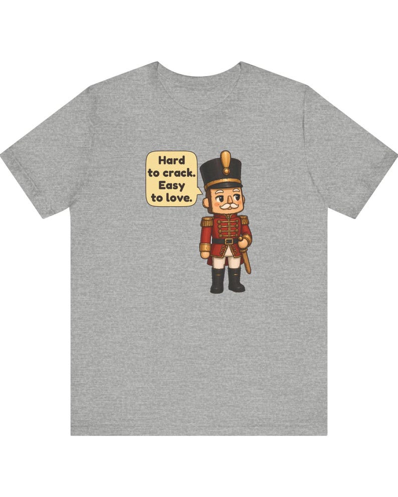 Pu&ograve; includere: Una t-shirt grigio melange con la grafica di un soldatino schiaccianoci in uniforme rossa con dettagli dorati. Accanto al soldatino c'&egrave; un fumetto con il testo: Hard to crack. Easy to love. Il disegno &egrave; centrato sul petto della maglietta.
