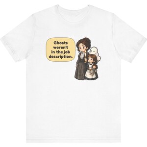 Op de afbeelding: Een wit t-shirt met een afbeelding van een persoon in een donkere jurk met een kaars, een kind met een pop en een klein spookje. Een tekstballon bevat de tekst: Ghosts weren't in the job description.