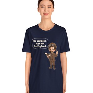 May include: A navy blue t-shirt featuring a cartoon illustration of an aviator with a mustache, goggles, and a brown suit holding a small airplane. A speech bubble contains the text: No compass. Just aim for England.