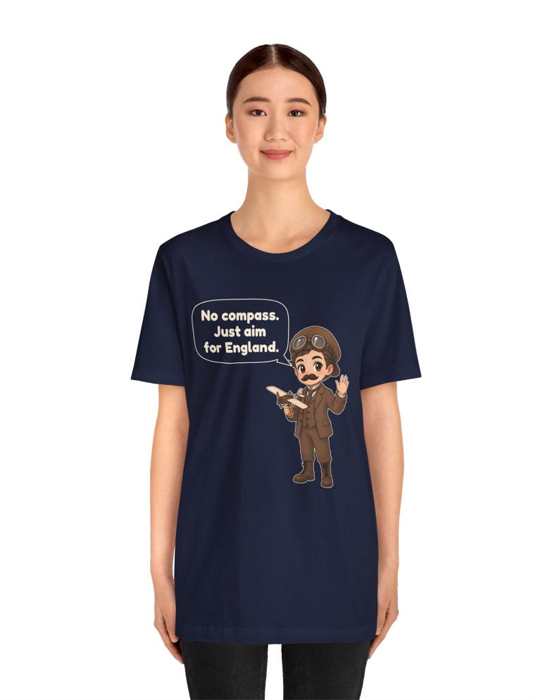 May include: A navy blue t-shirt featuring a cartoon illustration of an aviator with a mustache, goggles, and a brown suit holding a small airplane. A speech bubble contains the text: No compass. Just aim for England.